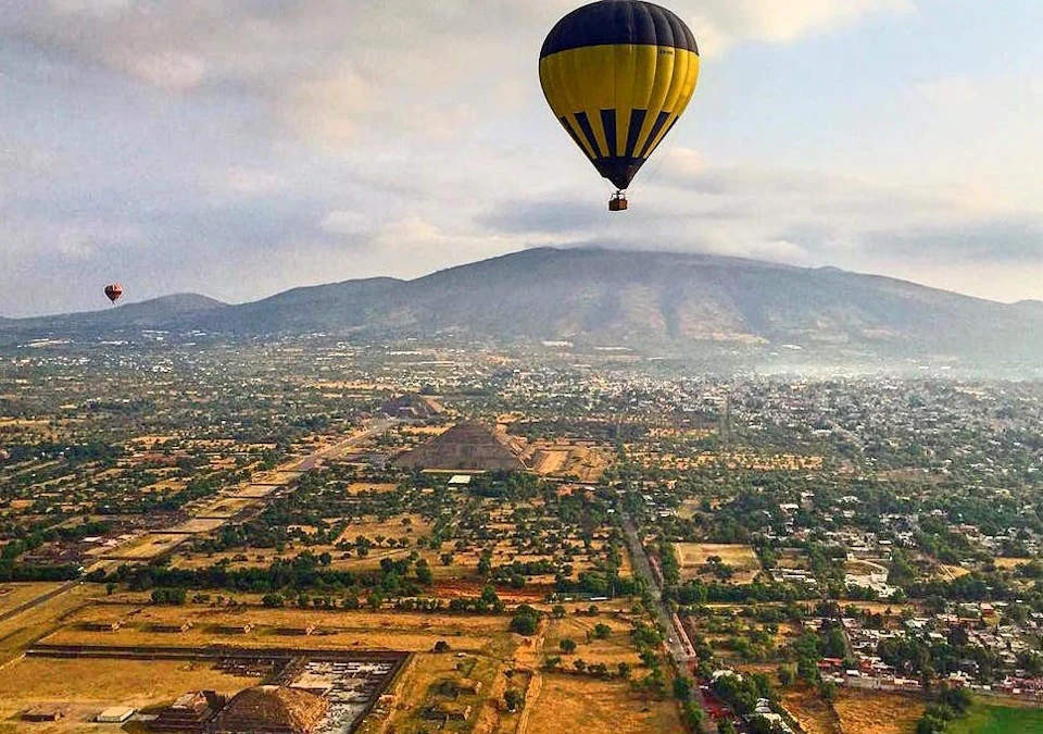 A yellow and black hot air balloon floats over a sprawling, green and brown landscape with mountains in the background.