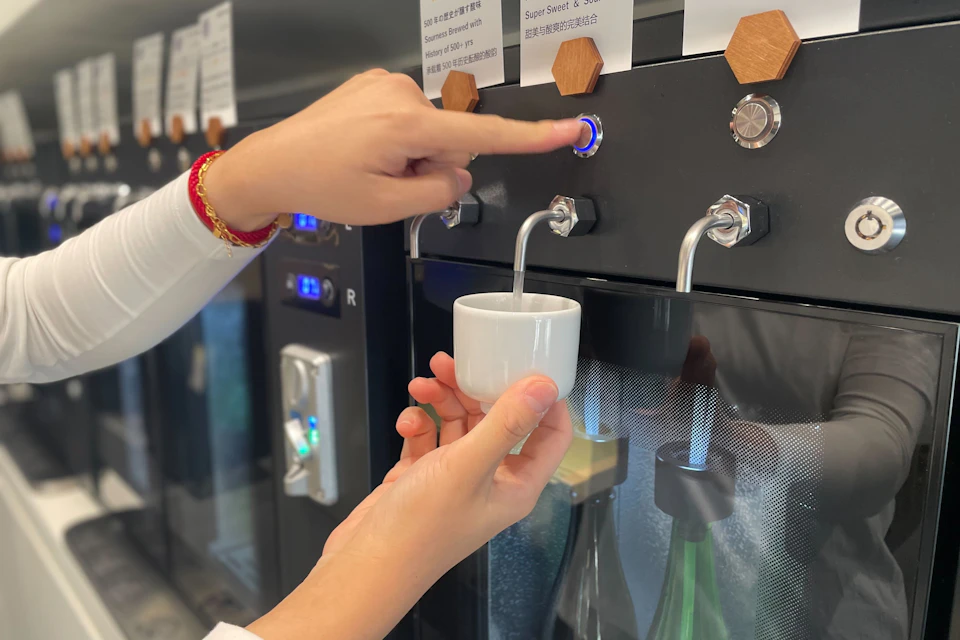 A person dispenses a beverage into a white cup from a machine, pressing a blue-lit button. Plastic bottles are partially visible below.