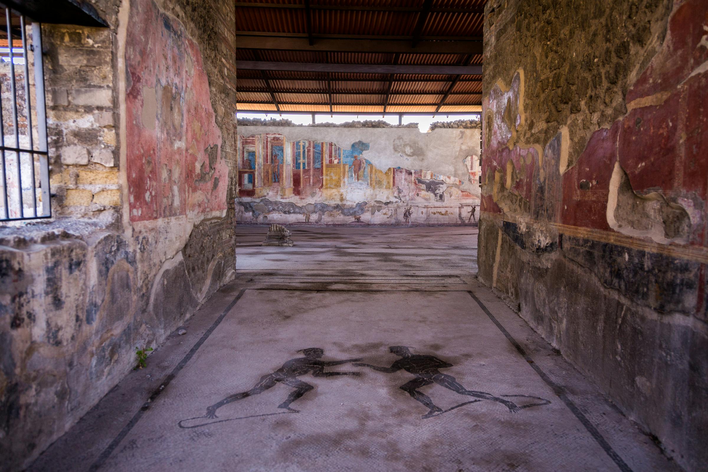 古代遗址，墙壁上有褪色的壁画，地板上有两个人物的马赛克，上面有一个木屋顶。