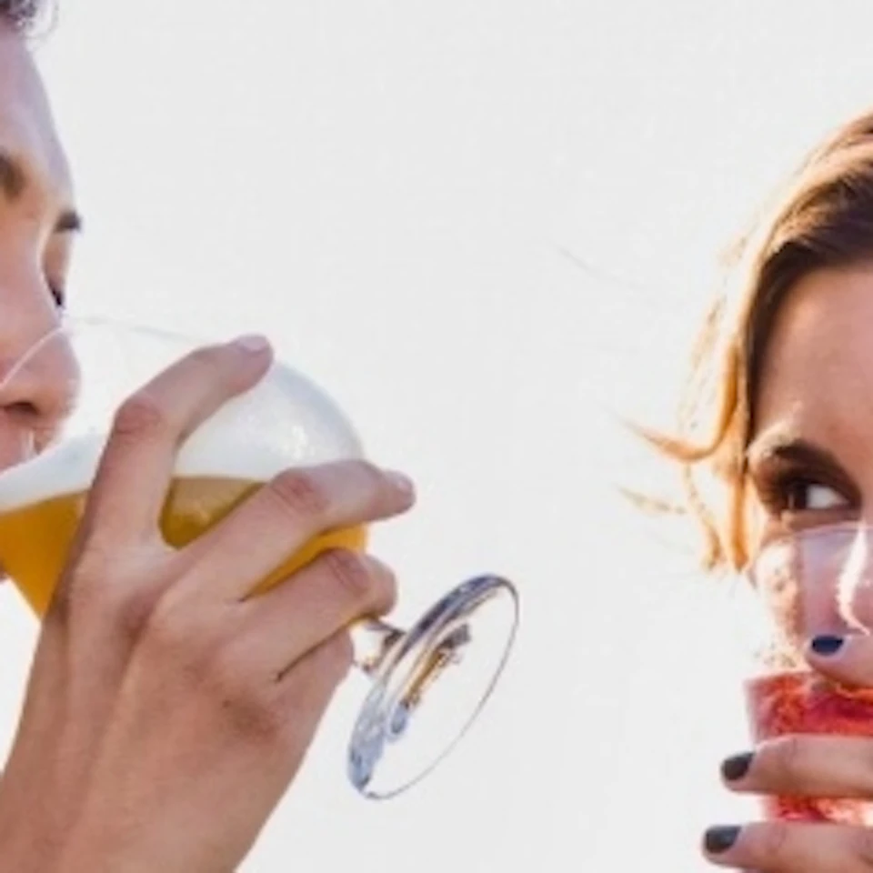 Two women in dresses on a boat deck, one drinking beer and the other holding a glass of rosé wine, with the ocean in the background.