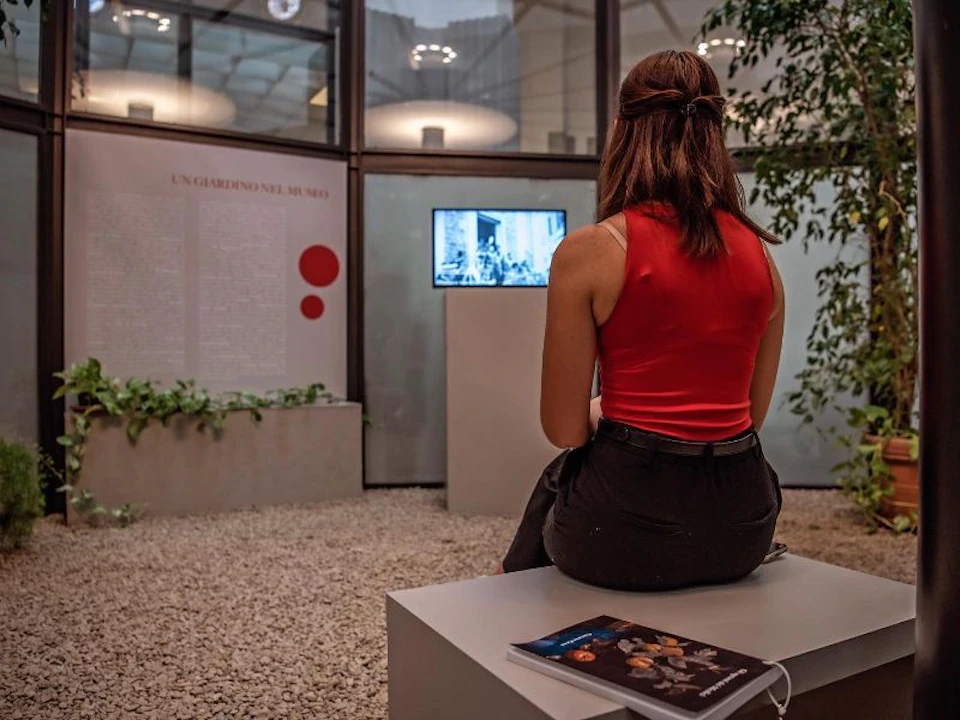 A woman in a red top sits on a bench in a modern museum, viewing a black-and-white display.