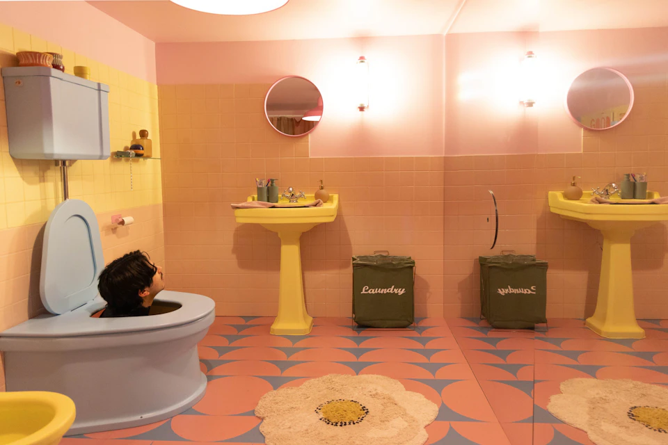 A retro-styled bathroom with pastel colors, a yellow sink, laundry bins, and a person partially seen in a bathtub.