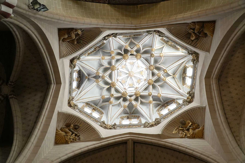 A decorative, intricate ceiling with an octagonal design, featuring gold accents and sculpted figures in a cathedral.