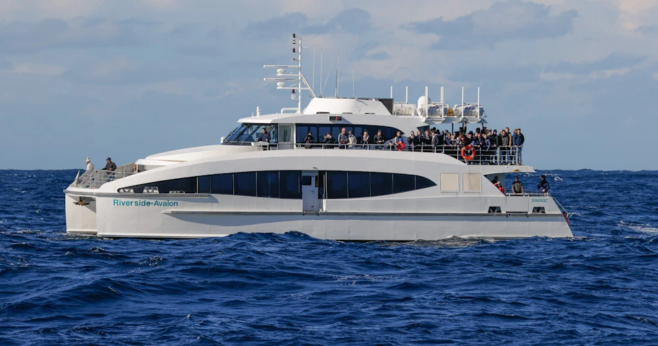 A white ferry with two decks crowded with passengers sails on a blue ocean under a partially cloudy sky.