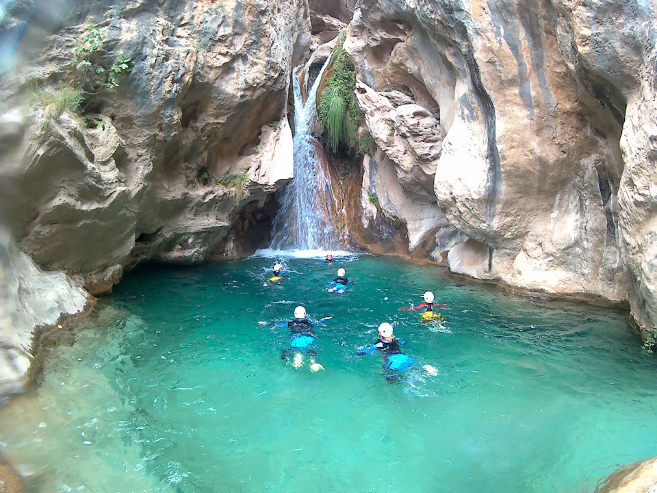 Five people wearing wetsuits and helmets swim towards a small waterfall in a rocky canyon with clear turquoise water.