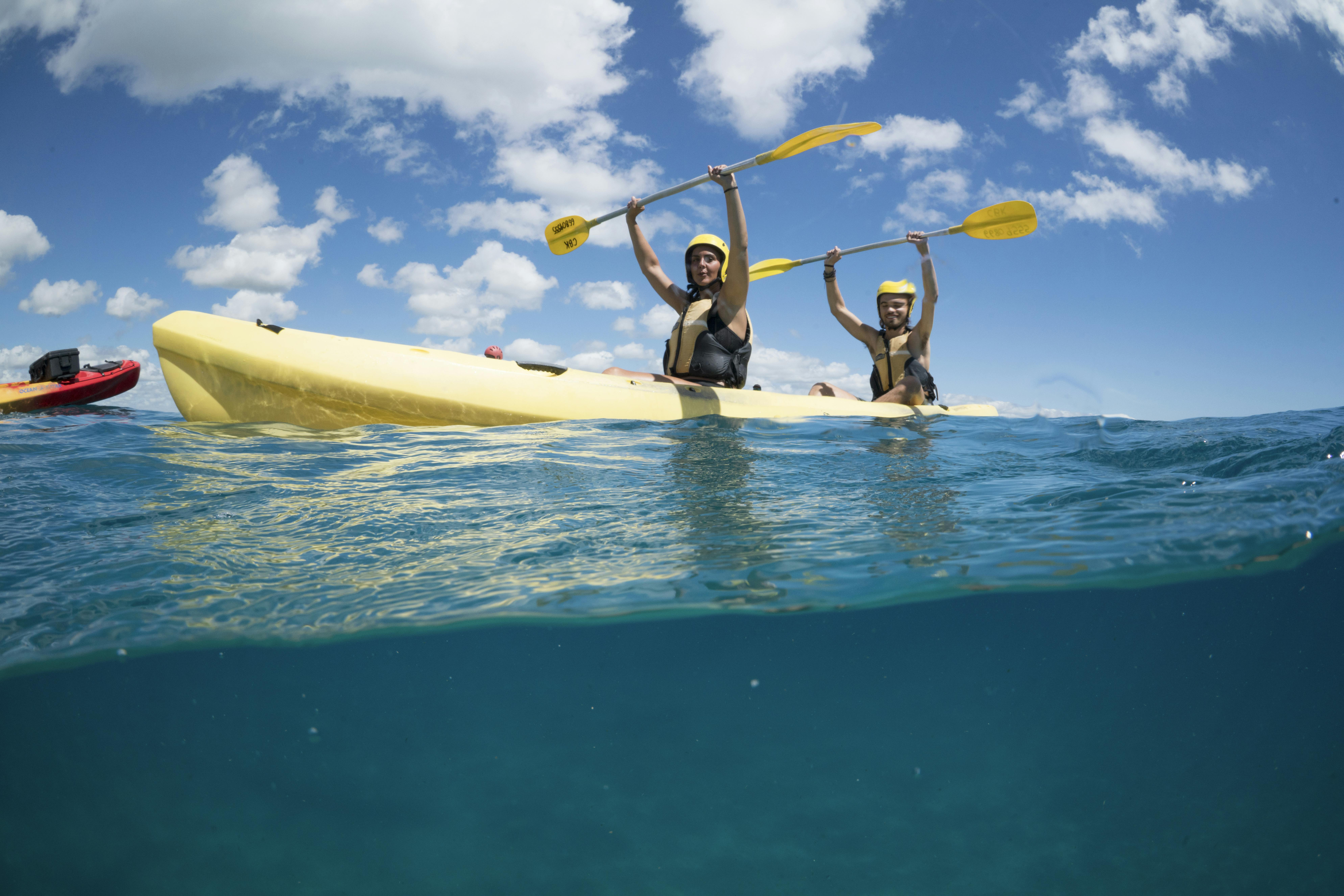 Two people in life vests hold paddles above their heads while sitting in a yellow kayak on blue water under a partly cloudy sky.