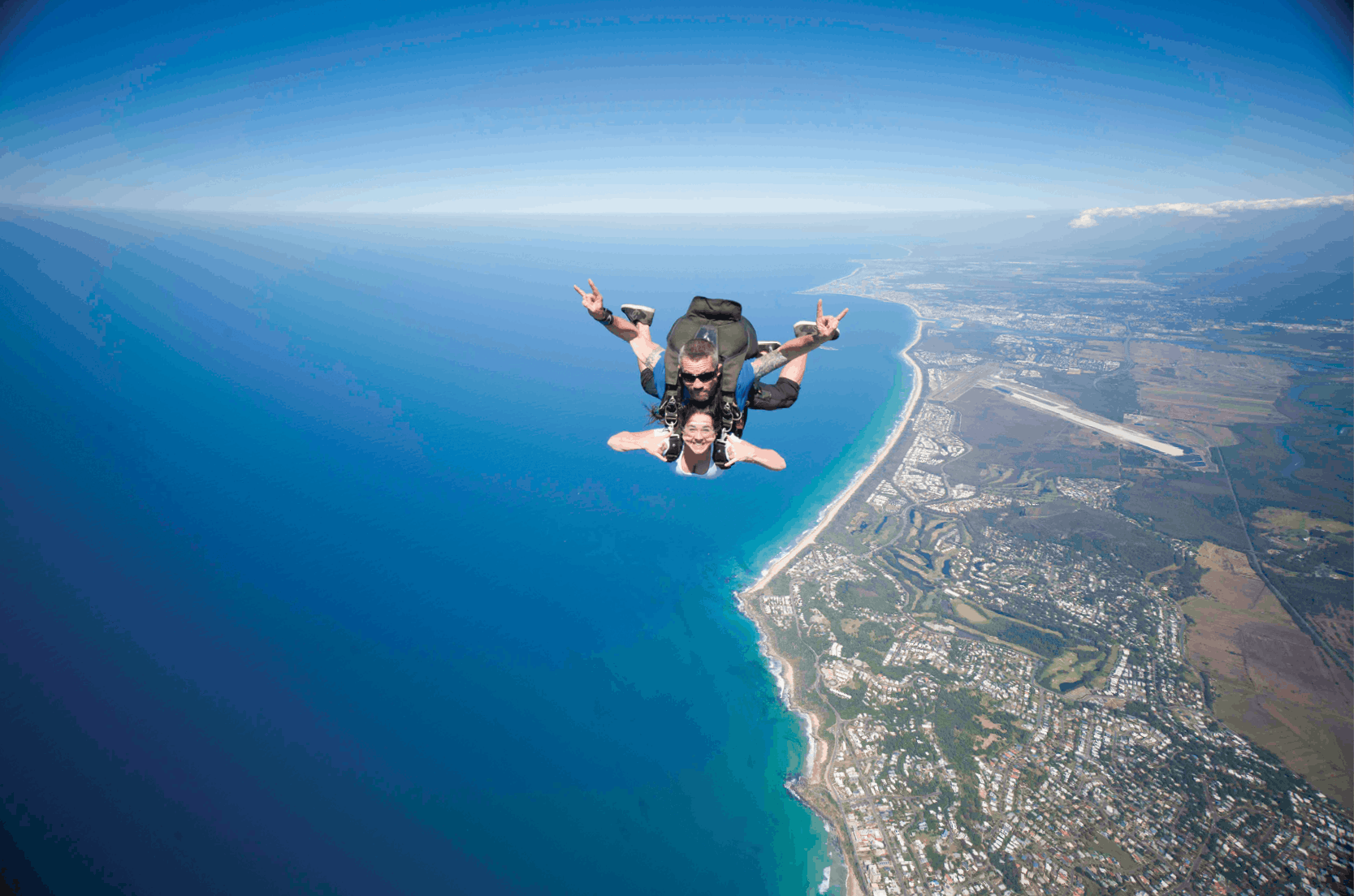 Two people tandem skydiving over a coastal landscape with sprawling city, blue ocean, and clear sky in the background.