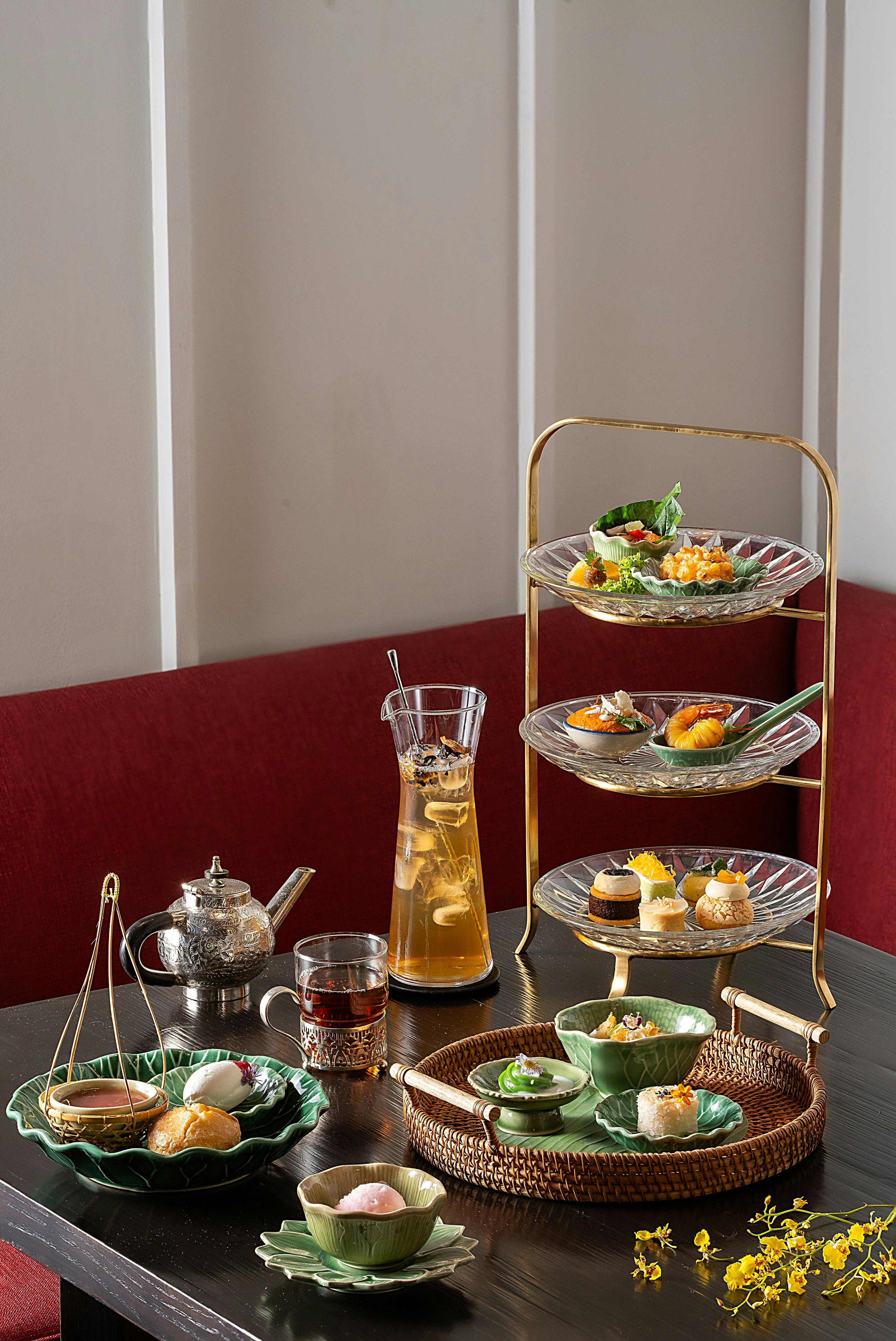A table set with a tiered tray of assorted foods, a teapot, glasses with drinks, and plates with small dishes, against a red booth.