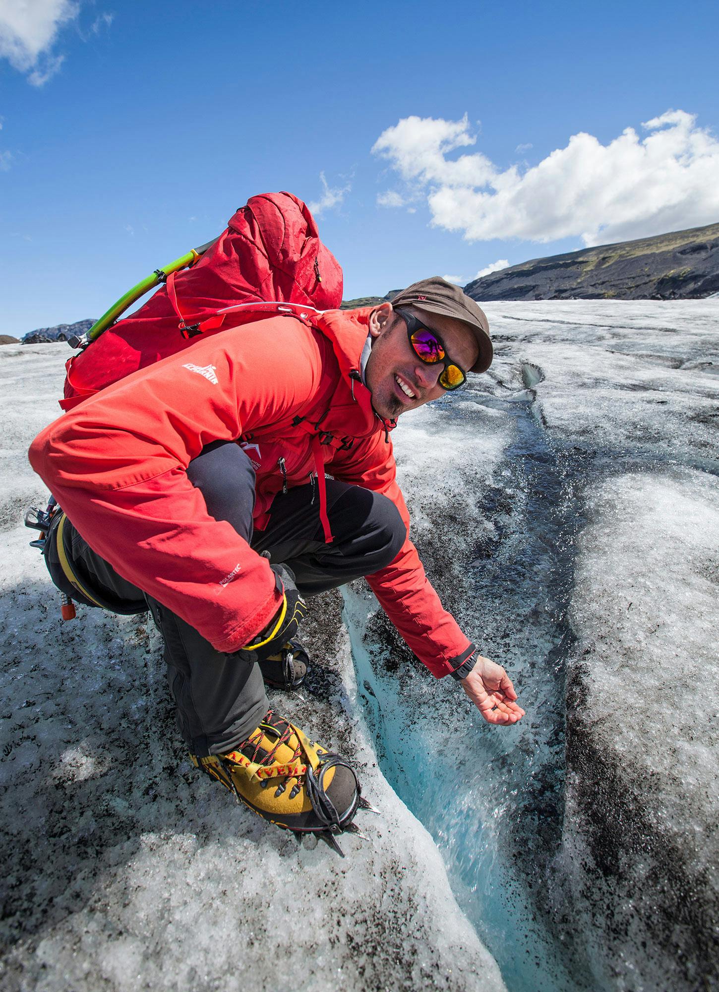 Man in red jacket and backpack crouching on icy terrain, wearing sunglasses and crampons, scooping water from a crevice. Clear sky above.