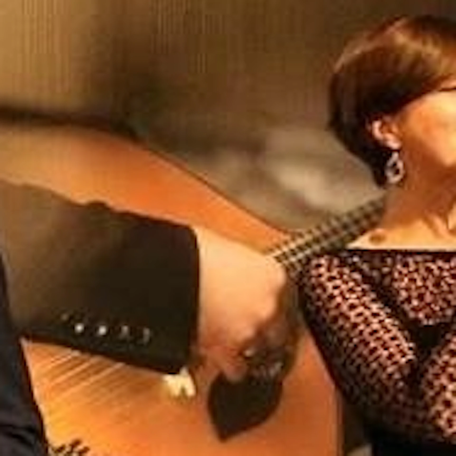 A woman sings on stage next to a man in a suit, with an audience watching in a rustic room adorned with ceramic vases.
