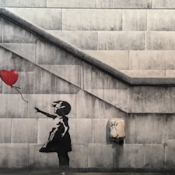 Graffiti av en flicka som sträcker sig efter en röd hjärtformad ballong på en betongvägg med texten "DET FINNS ALLTID HOPP".