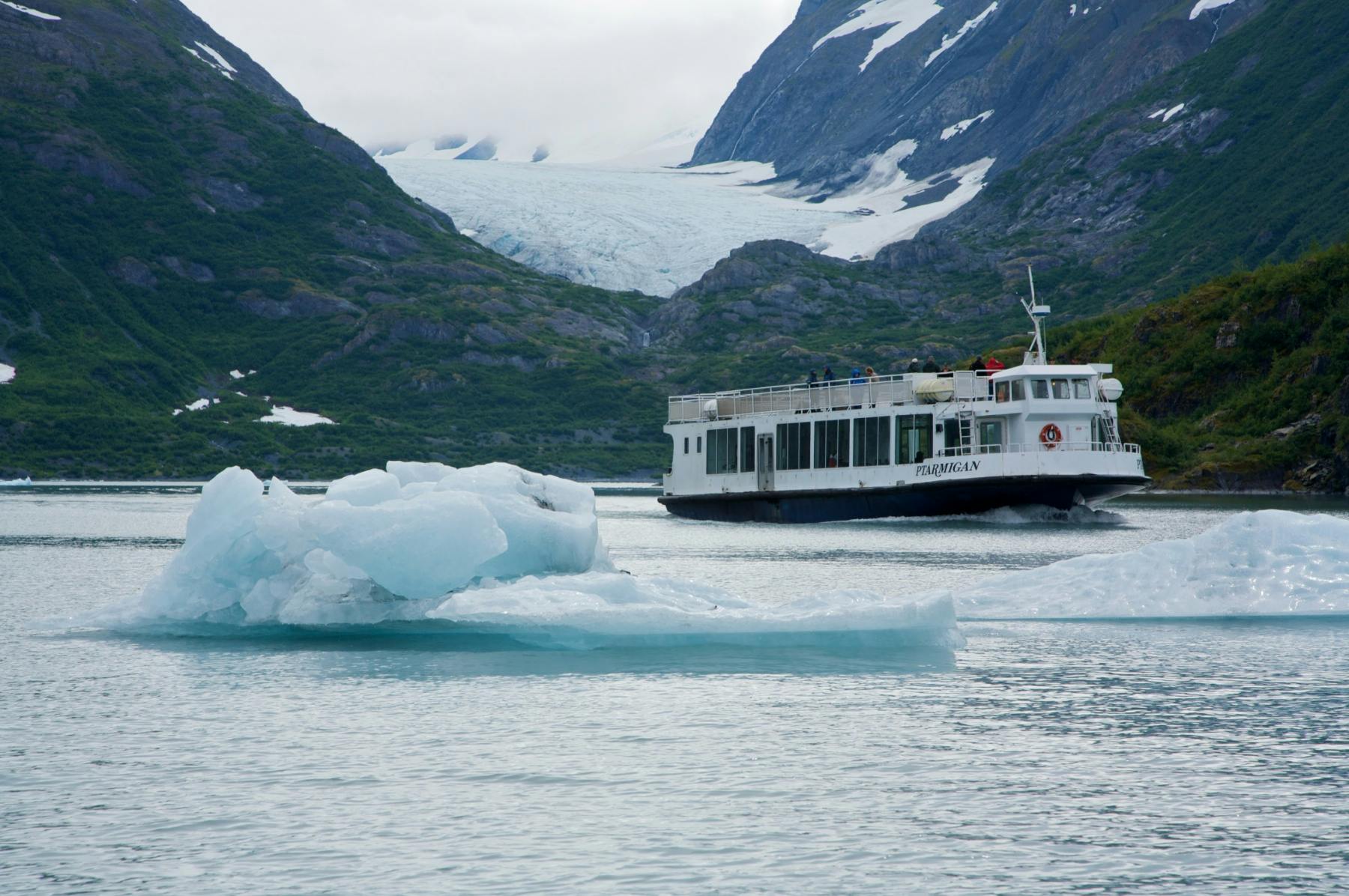 A tour boat navigates near icebergs on a lake surrounded by green mountains and a glacier in the background under cloudy skies.