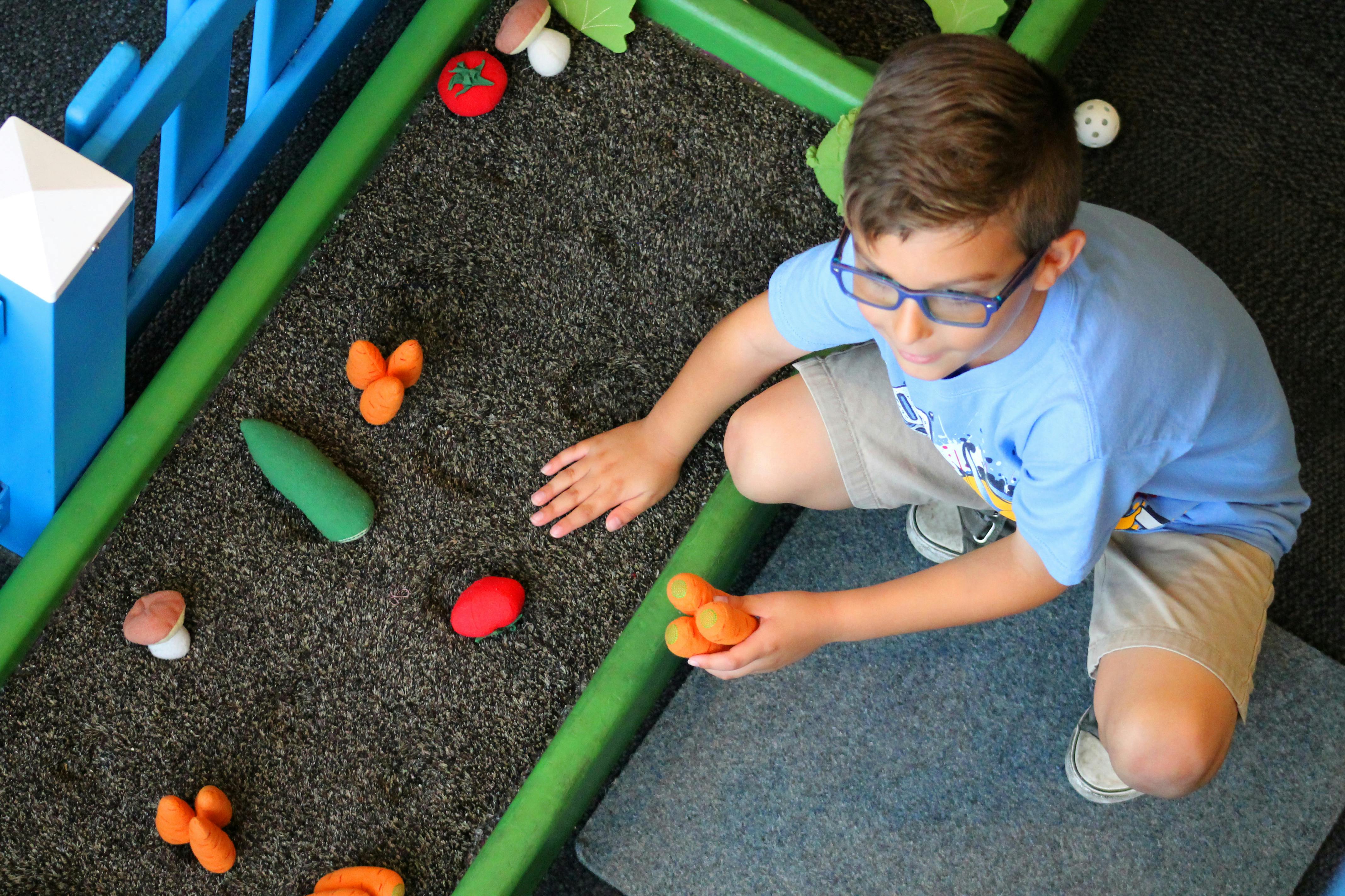 Mladý chlapec v brýlích a modré košili drží oranžové kuličky a hraje si na koberci se zelenými okraji a malými hračkami.
