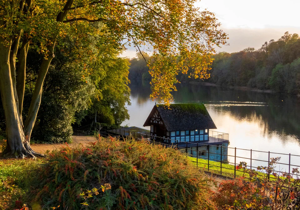 Scenic lakeside view with foliage, a timber-framed boathouse on the shore, and trees reflecting on the calm water at sunset.