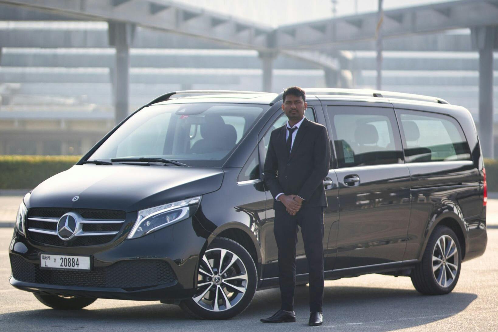 En man i svart kostym står bredvid en svart minibuss på en parkeringsplats.