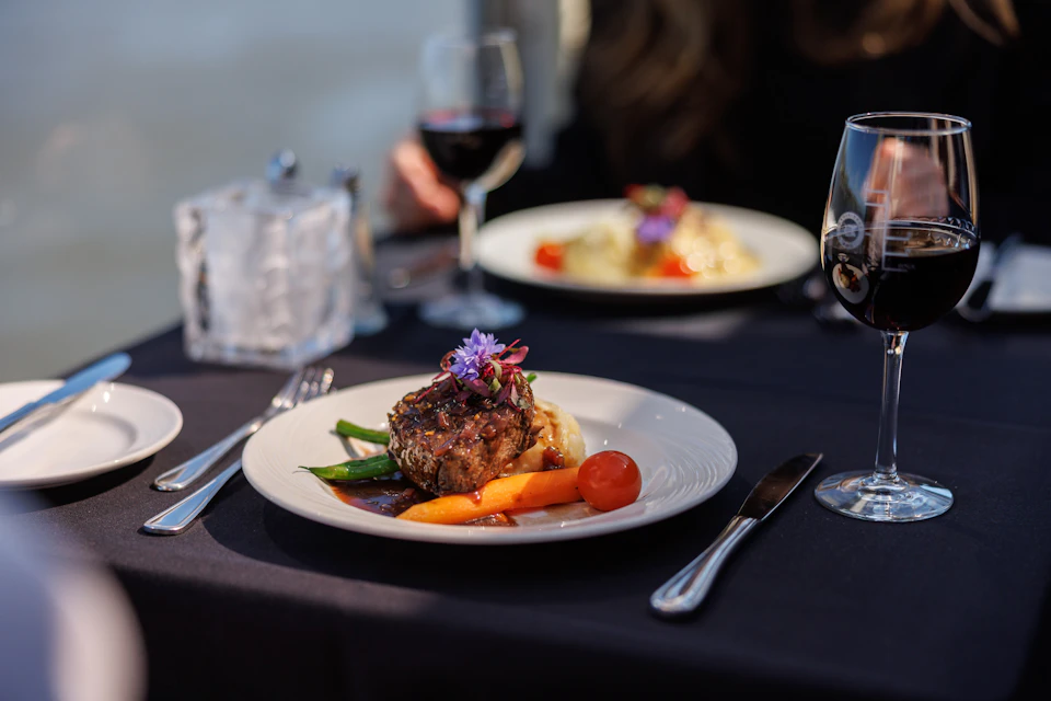 A plate with a gourmet meal including meat, vegetables, and a garnish on a table with wine glasses and another plate visible.