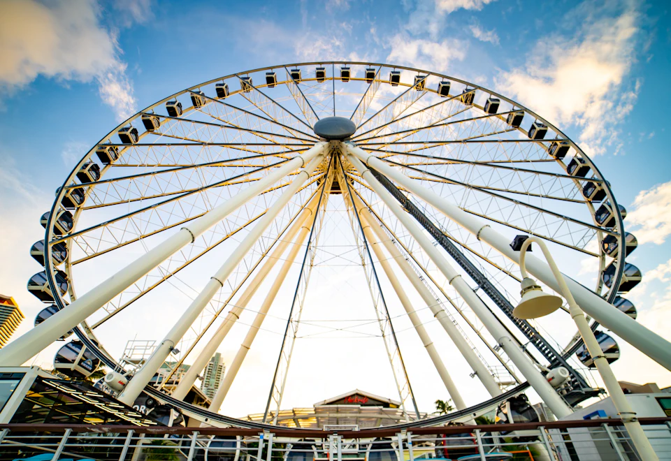Low angle view of a large Ferris wheel against a blue sky with scattered clouds. The base of the wheel has a small building.