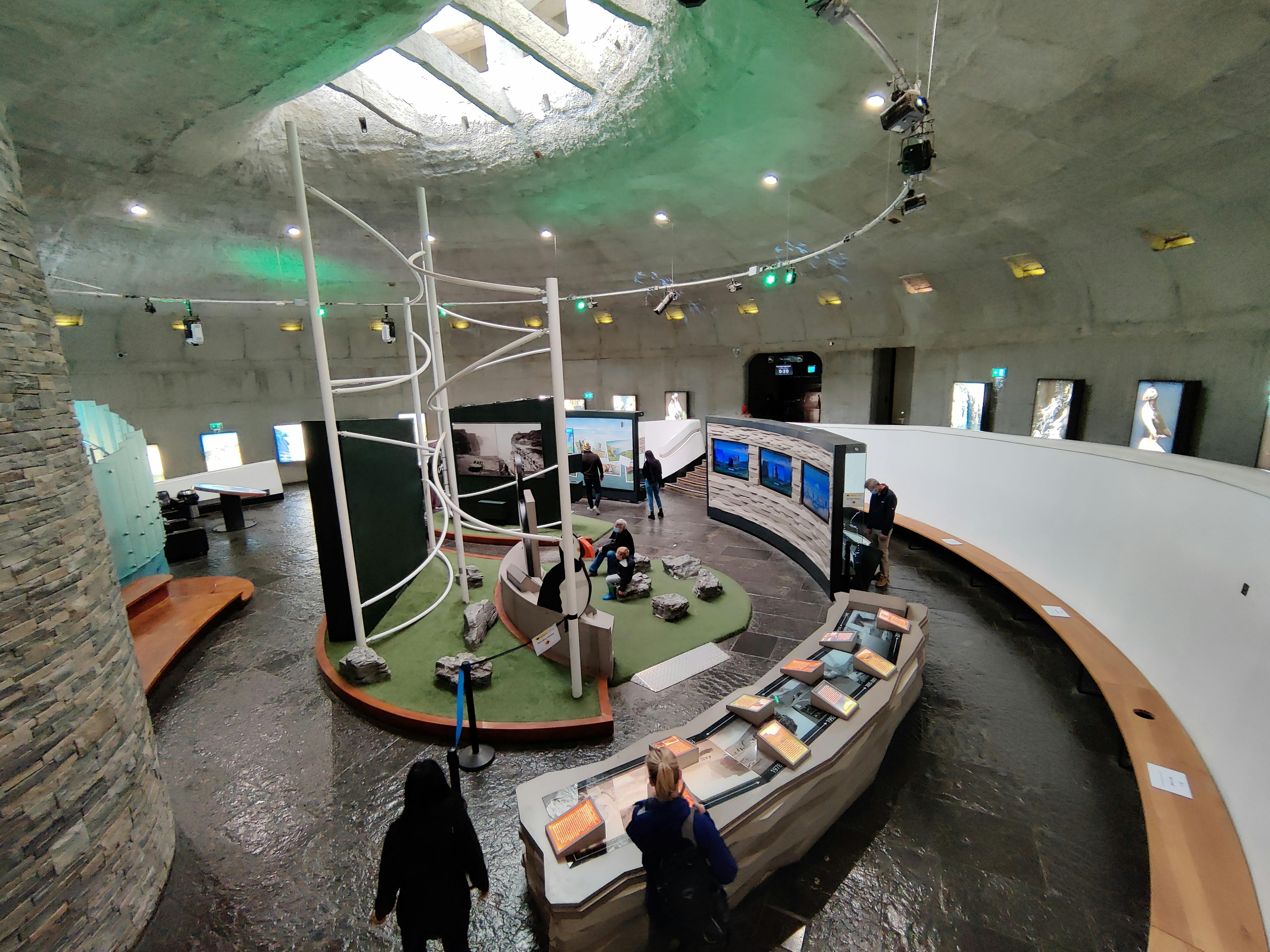 Visitors in a museum exhibit featuring geology displays, interactive screens, and a spiral structure under a circular skylight.