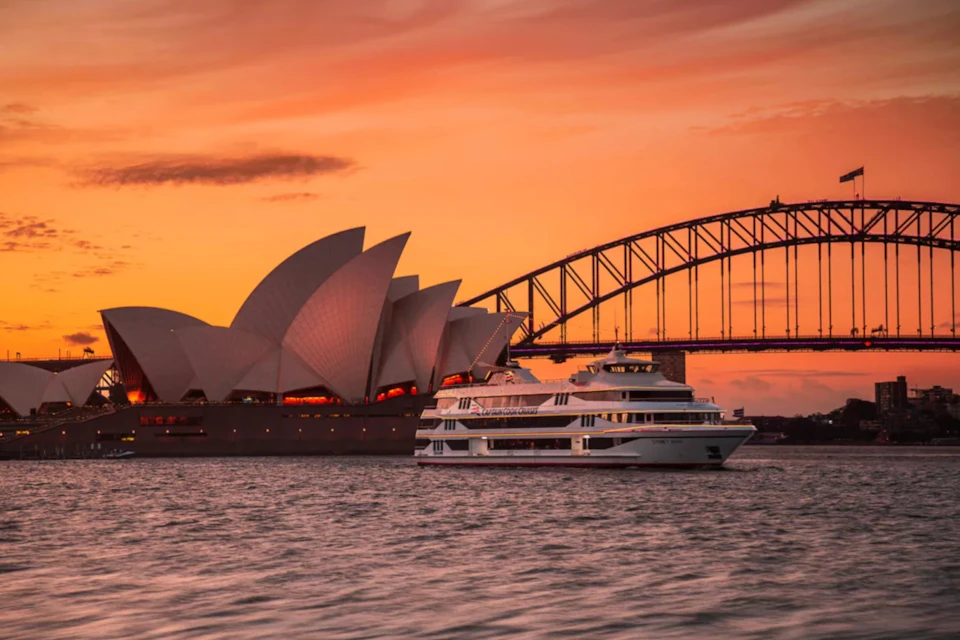 A white boat sails past the Sydney Opera House and Sydney Harbour Bridge at sunset under an orange sky.