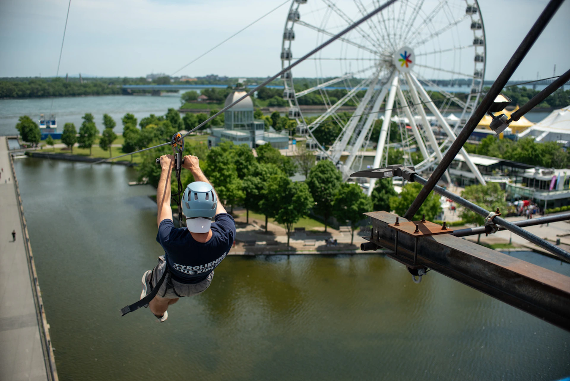 Person ziplining over a body of water with a Ferris wheel and green trees in the background.