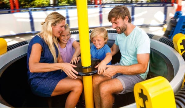 Eine vierköpfige Familie sitzt zusammen auf einem Fahrgeschäft, lächelt und hält sich an einer gelben Stange in der Mitte fest.