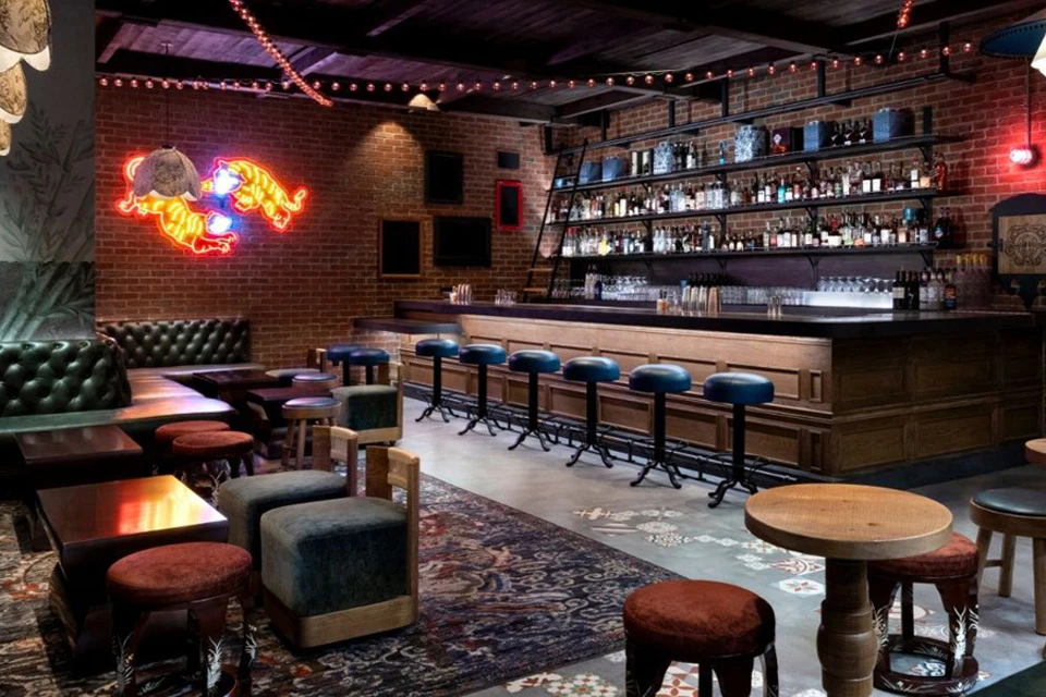 Cozy bar interior with a wooden counter, barstools, shelves of bottles, and a neon sign on a brick wall.