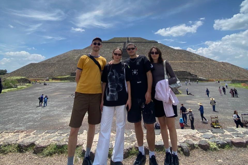 Four people standing in front of a large ancient pyramid under a partly cloudy sky.