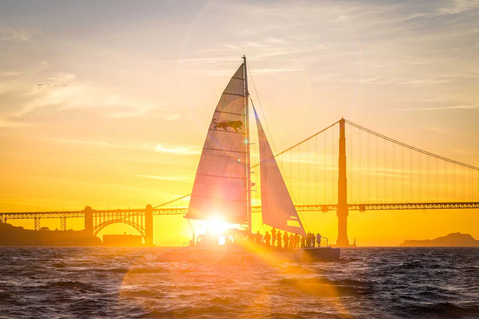 A sailboat with people aboard sails at sunset near a large suspension bridge. Sunlight creates a warm glow and lens flare effects.