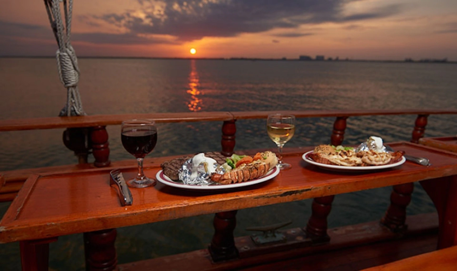 Two plates of food and two glasses of wine on a wooden railing with a sunset over a calm body of water in the background.
