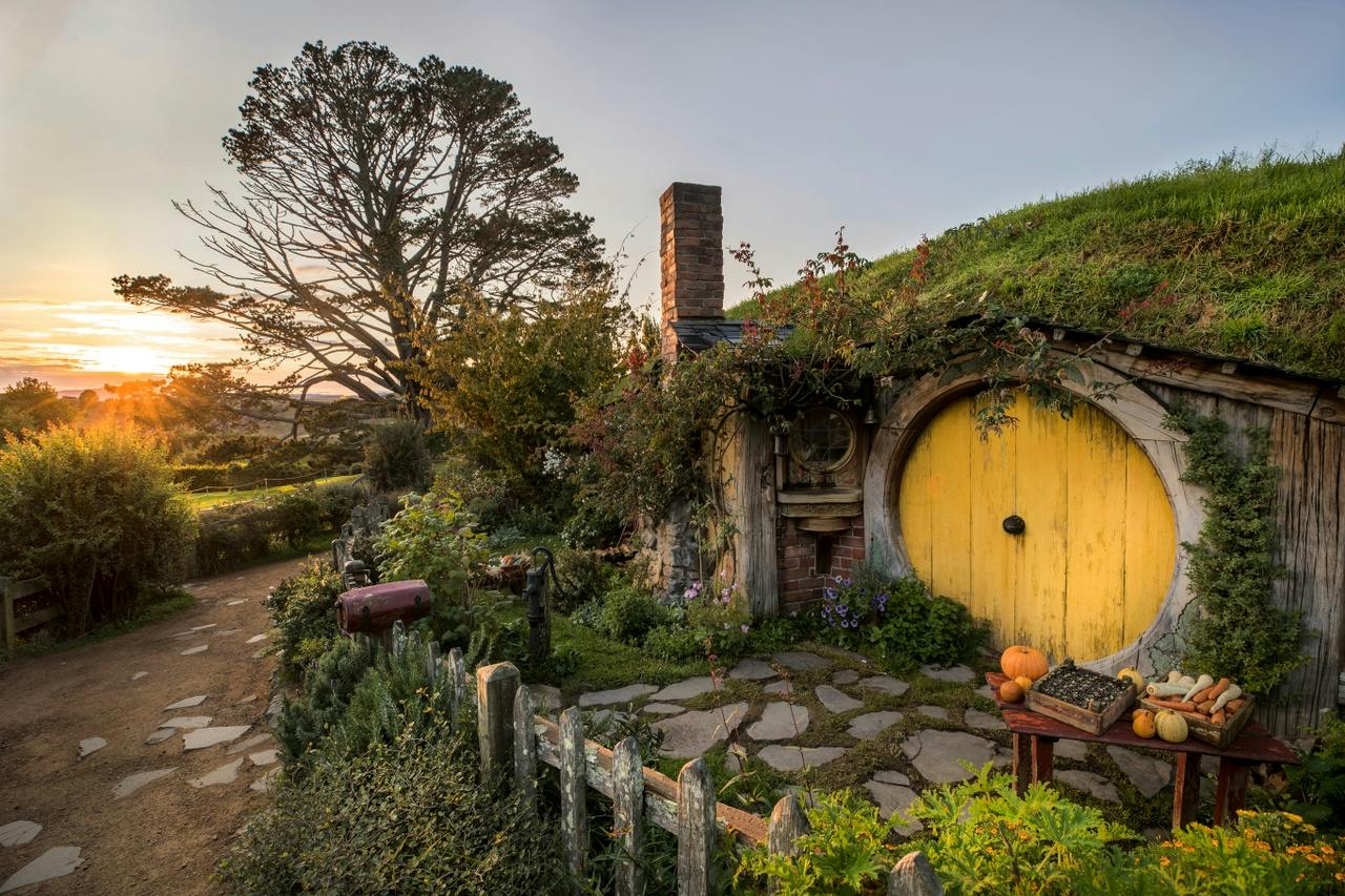 A cozy hobbit-style house with a yellow round door, a grassy roof, and a rustic garden at sunset, surrounded by lush greenery.