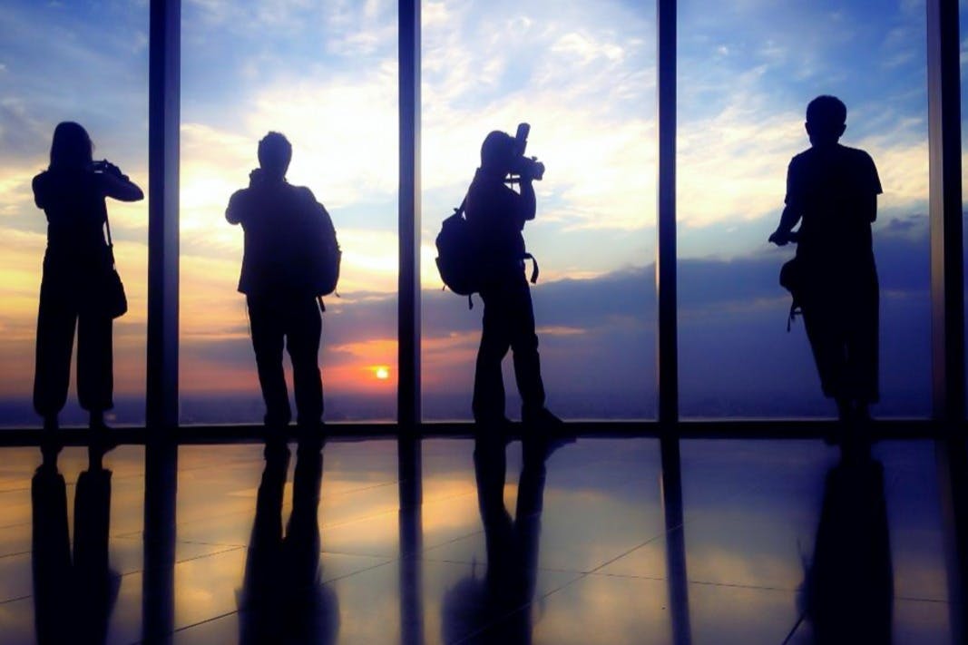 Silhouettes of three people with backpacks by a large window capturing a sunset over the horizon.