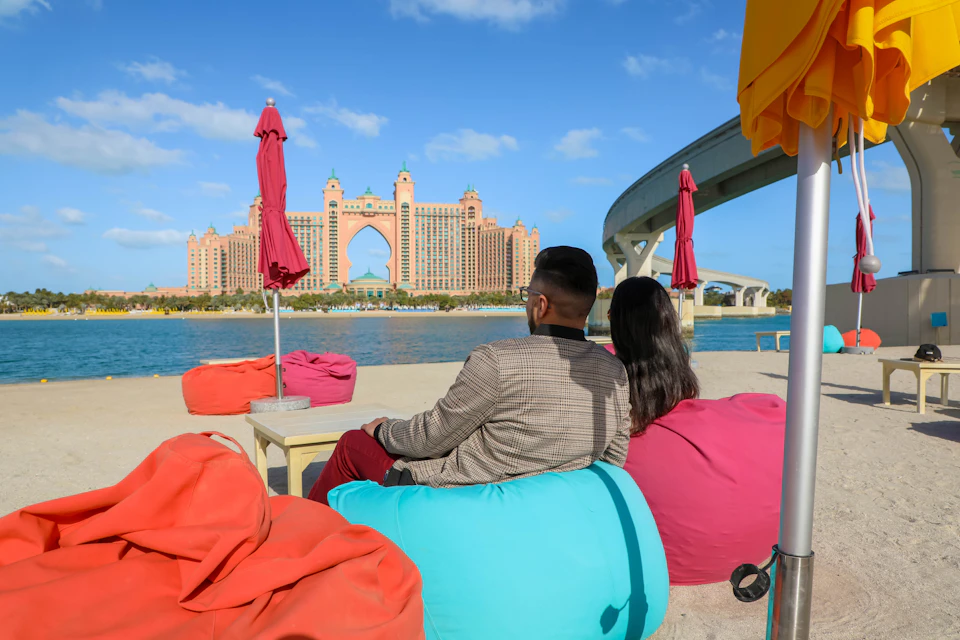 A man and woman sit on colorful bean bags near the water, with a large, ornate hotel in the background and some red umbrellas.