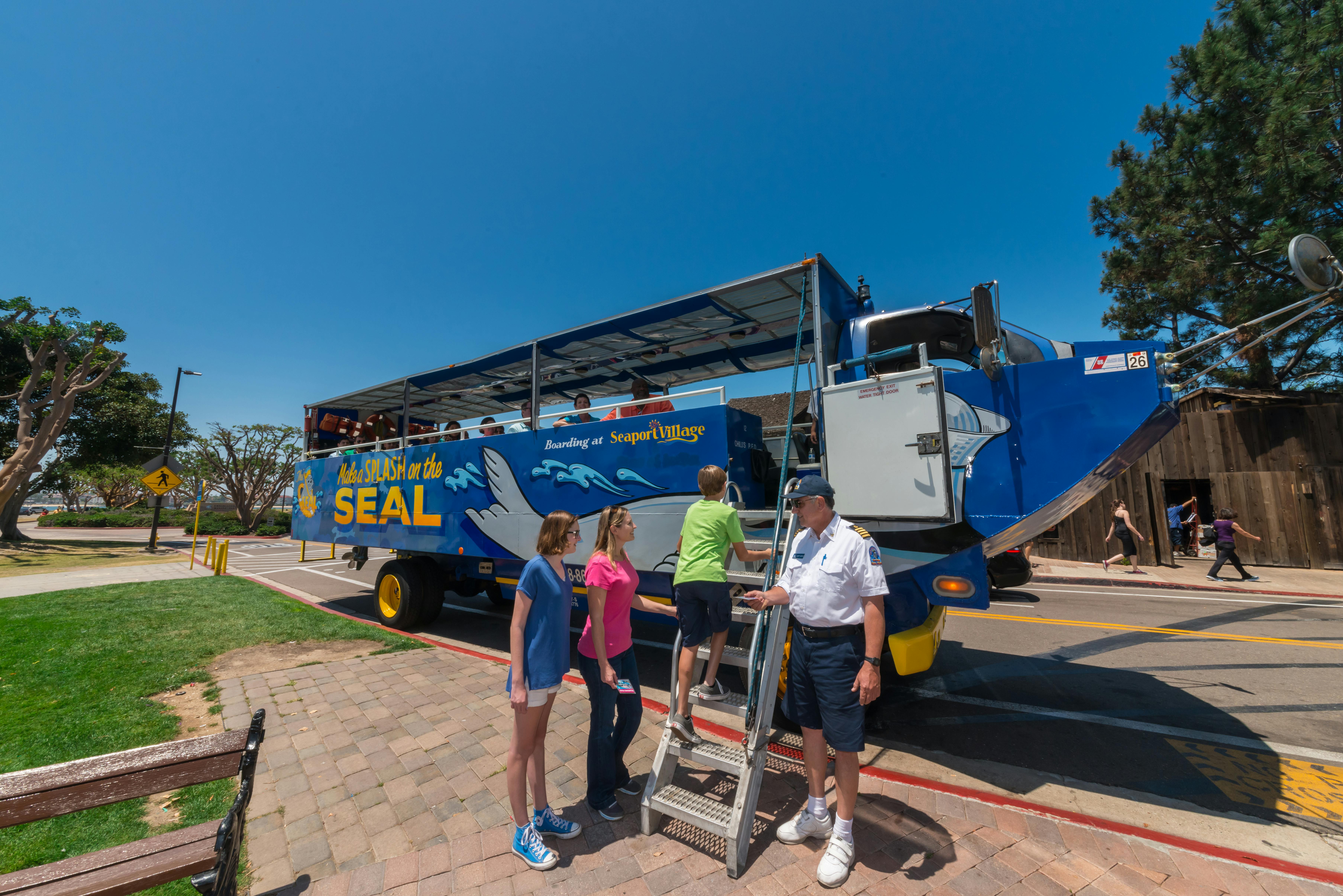People boarding a blue amphibious tour bus named "SEAL" with a driver in uniform assisting them under a clear blue sky.