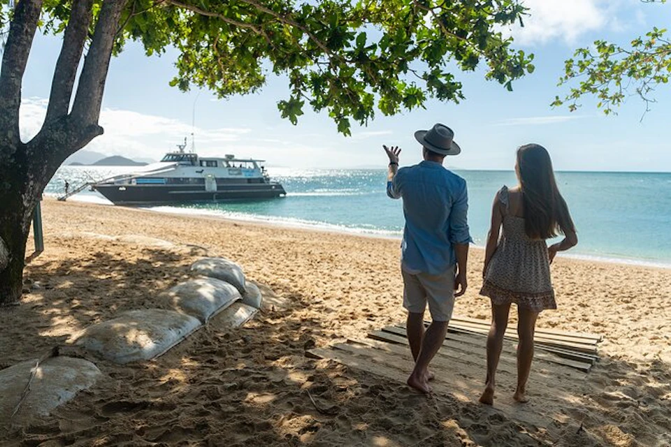 A man and woman stand on a sandy beach under tree shade, watching a boat on the water.