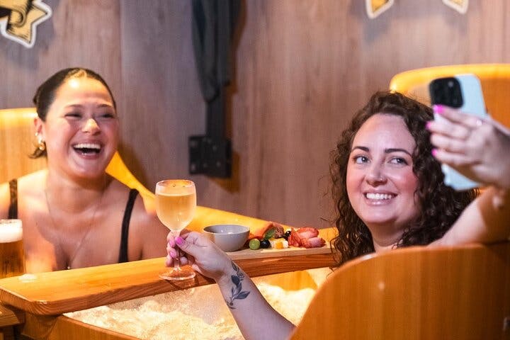 Δύο γυναίκες χαμογελούν και χαλαρώνουν σε ένα ξύλινο τζακούζι, η μία κρατώντας ένα ποτήρι κρασί. Στο παρασκήνιο είναι ορατός ένας μικρός δίσκος με σνακ.