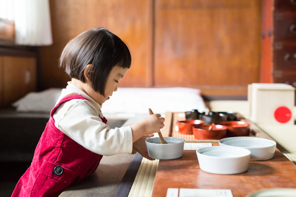 A young child wearing a red outfit sits and uses a spoon with white bowls and wooden elements in a traditional setting.