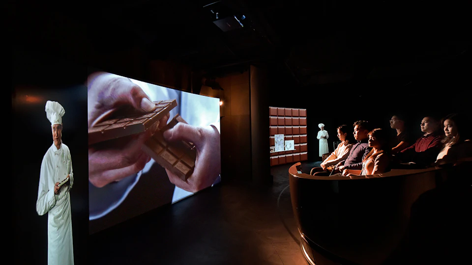 People seated in a dark room watching a large screen showing hands breaking a chocolate bar; an illuminated mannequin stands nearby.