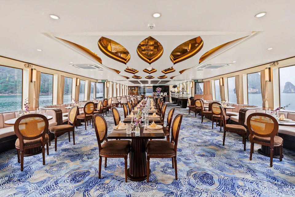 An elegant dining room with a blue swirling-pattern carpet, wooden tables, and brown upholstered chairs, featuring golden ceiling accents.