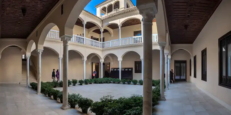 Une cour à deux étages avec des colonnes voûtées, des plantes en pot et des personnes marchant dans des couloirs. Le plafond est orné de motifs en bois complexes.