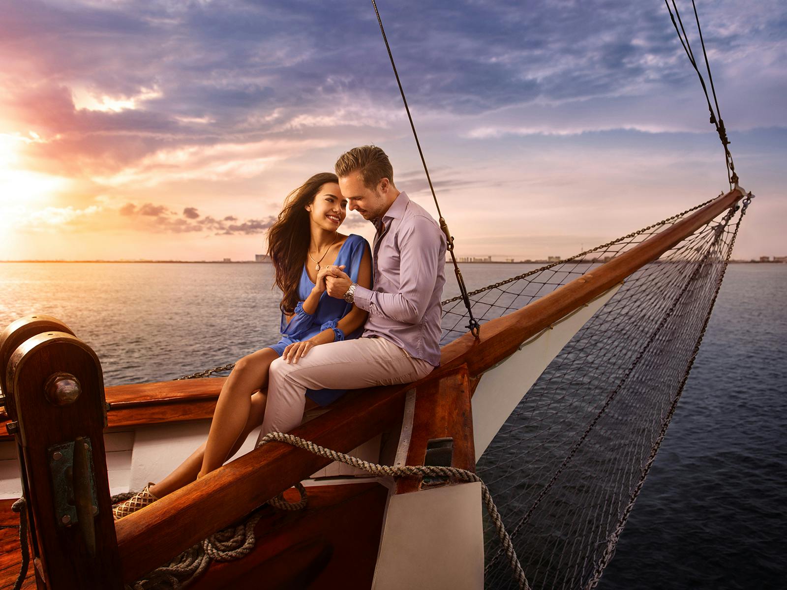 Una coppia siede a prua di una barca a vela in legno durante il tramonto, sorridendo e tenendosi per mano sullo sfondo dell'oceano.