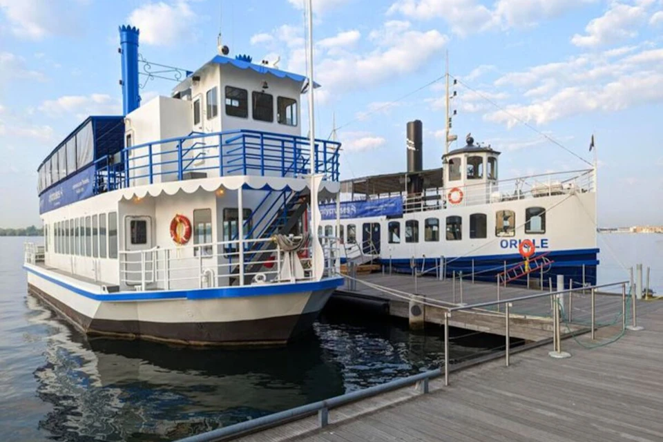 Two white and blue passenger boats docked at a pier, with a calm water surface and cloudy sky in the background.