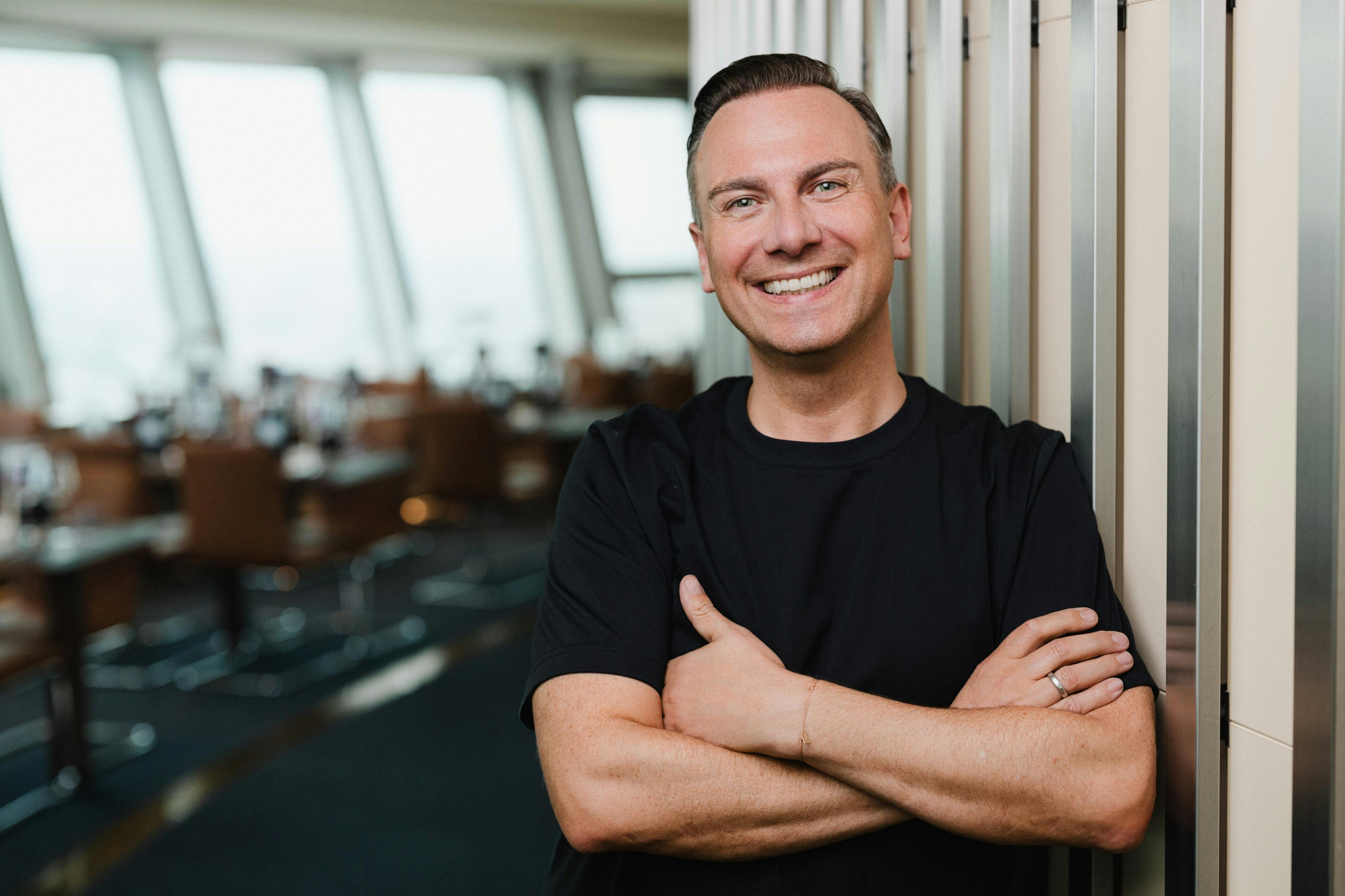 A smiling person in a black shirt standing with arms crossed in a modern indoor setting with large windows in the background.