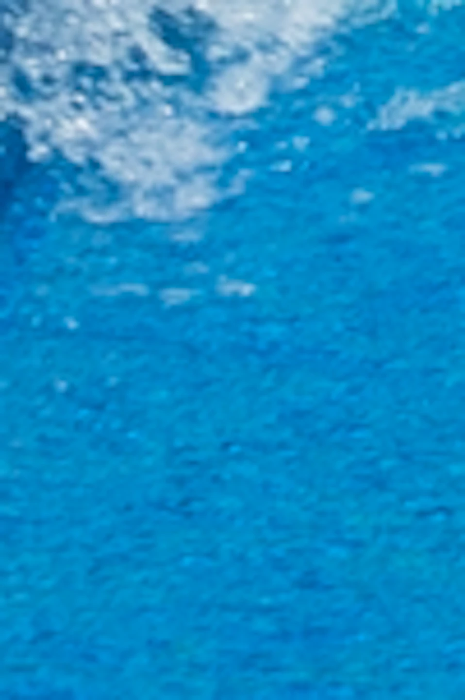People slide down colorful water slides on mats, splashing into a pool, with greenery and a covered structure in the background.