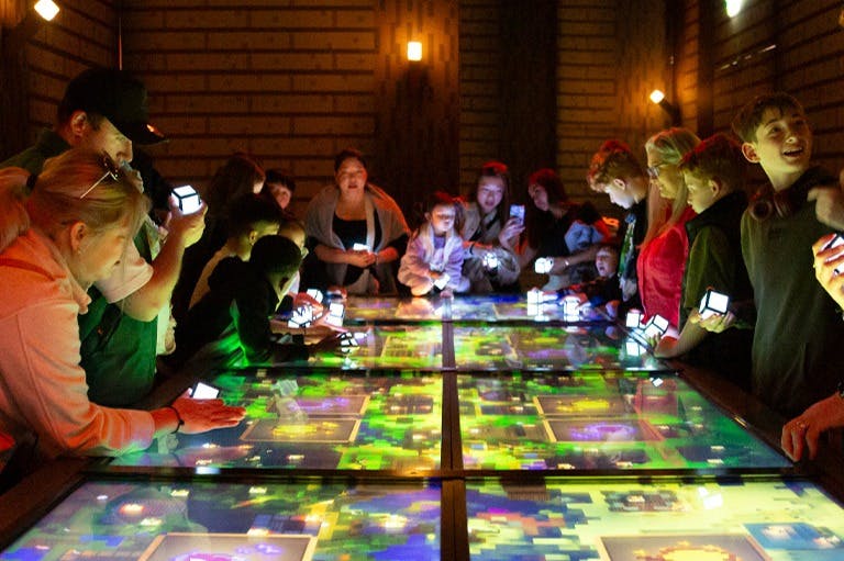 Lidé se shromáždili kolem osvětlených interaktivních stolů, zapojili se do displejů a drželi malé zářící kostky.