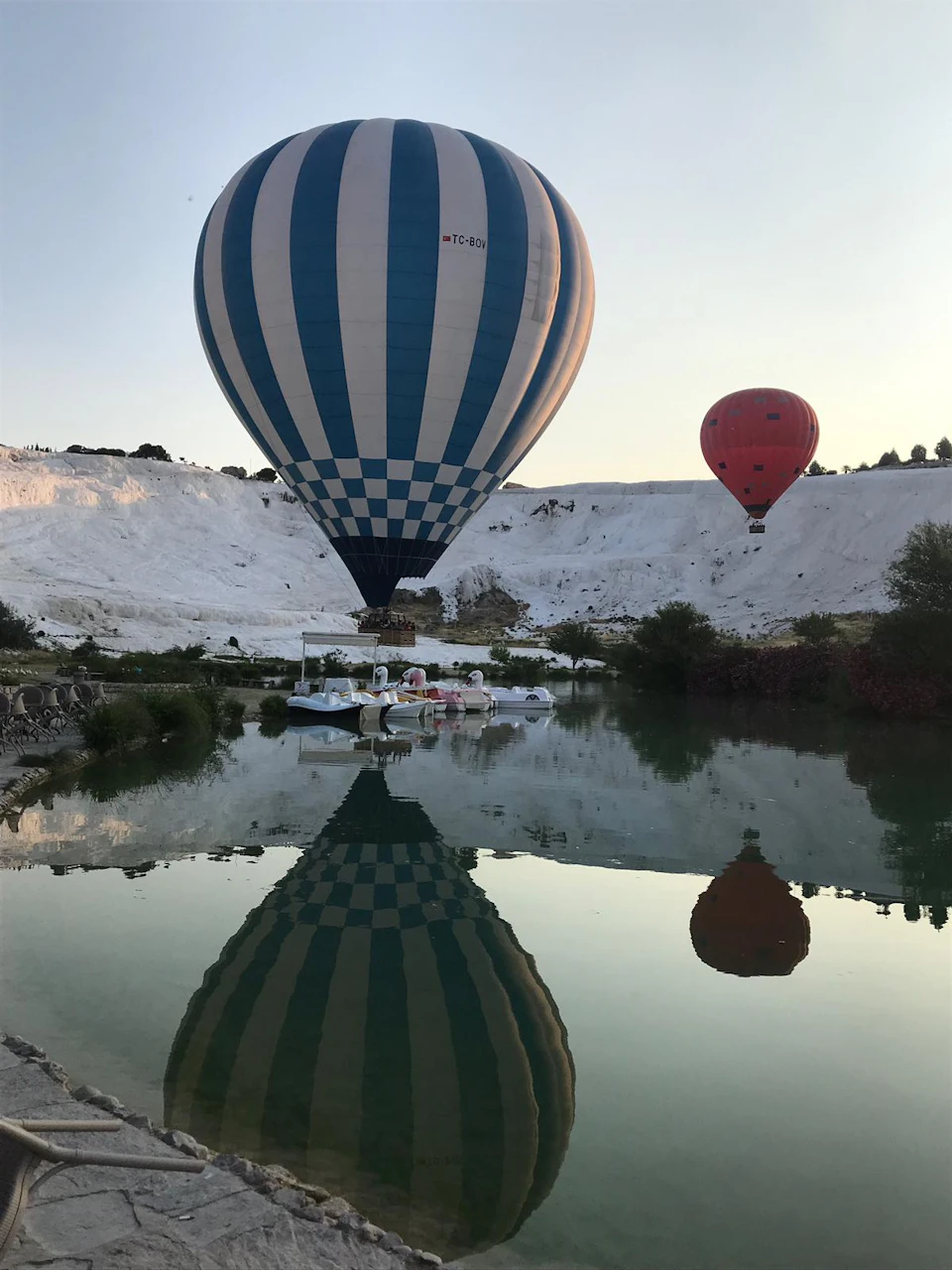 A blue-and-white hot air balloon with its reflection in a pond, with another red balloon in the background over white terrain.