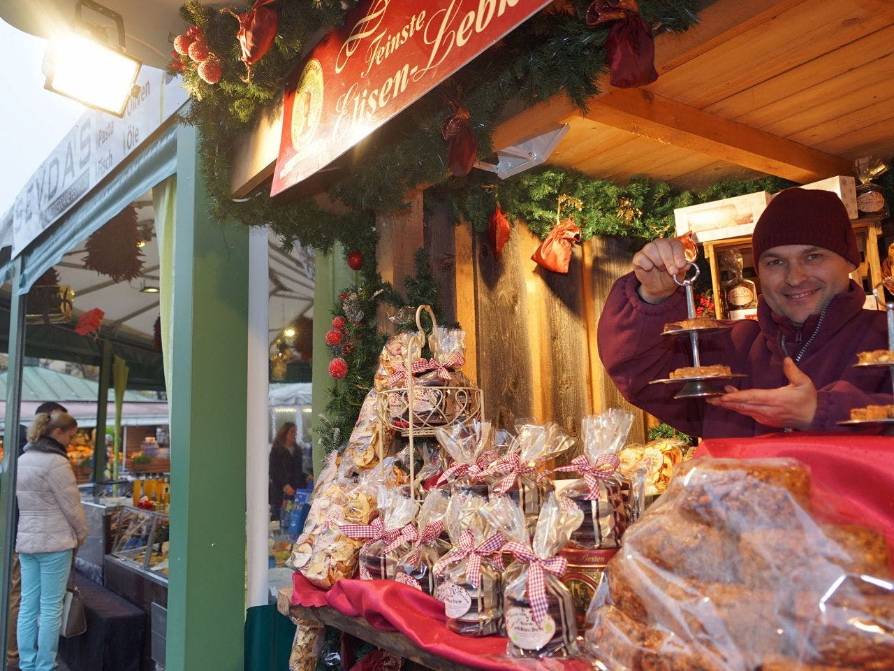 市场摊位上摆放着糕点，红色豆豆店的摊贩拿着分层的饼干托盘，陈列着成袋的烘焙食品。