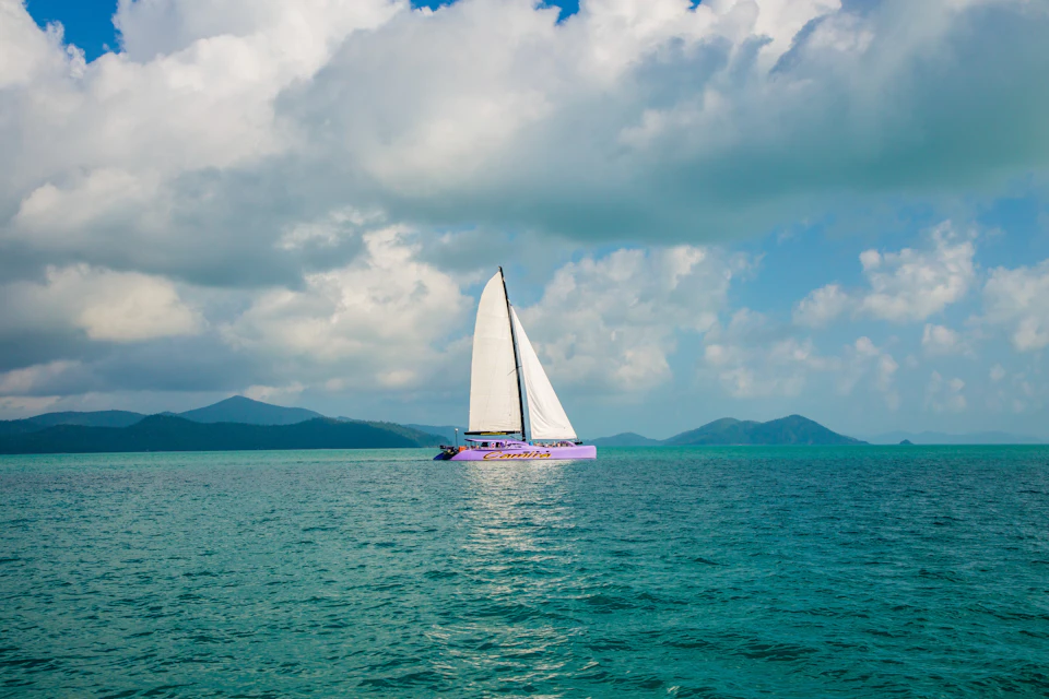A sailboat with a white sail and purple hull floats on calm turquoise waters with mountains and cloudy skies in the background.