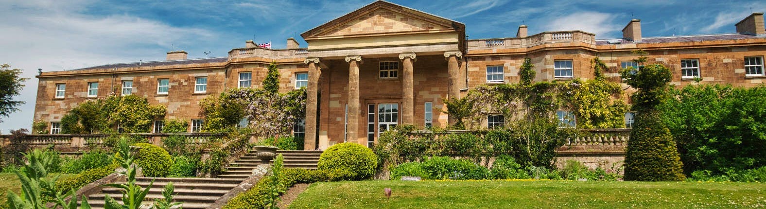Stor, historisk herrgård med en kolonnad ingång, stentrappor, grönska och välskötta buskar under en blå himmel.