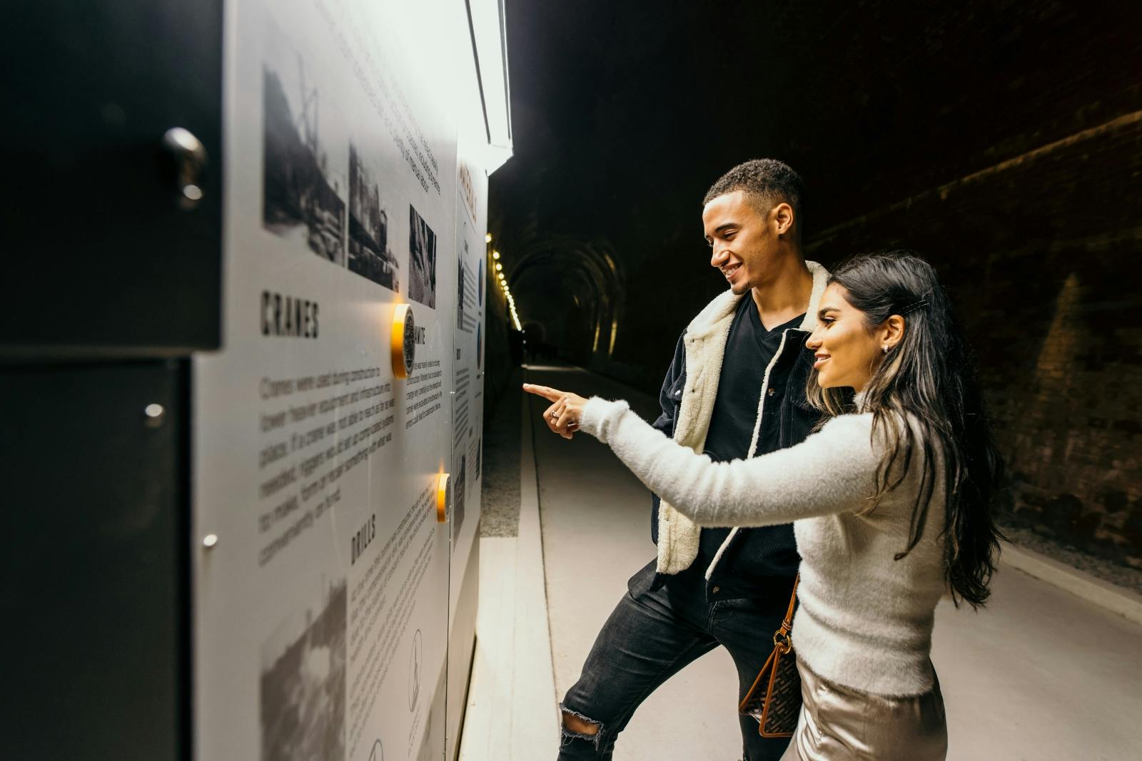 To personer interagerer med et informationsdisplay inde i en tunnel. Den ene peger på tekst, mens den anden ser på og smiler.