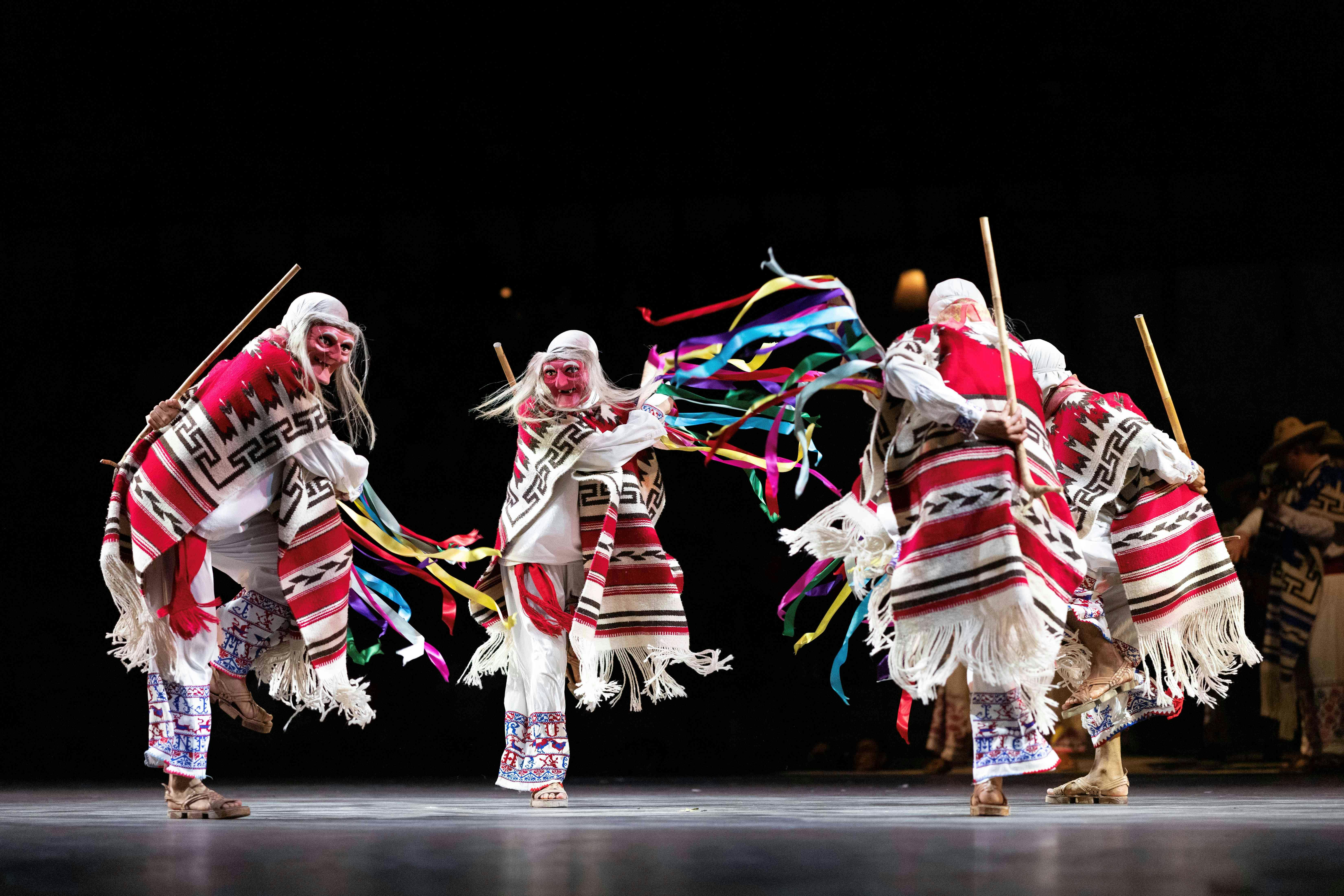 Performers in traditional attire with colorful ribbons dance on a stage under bright lights.